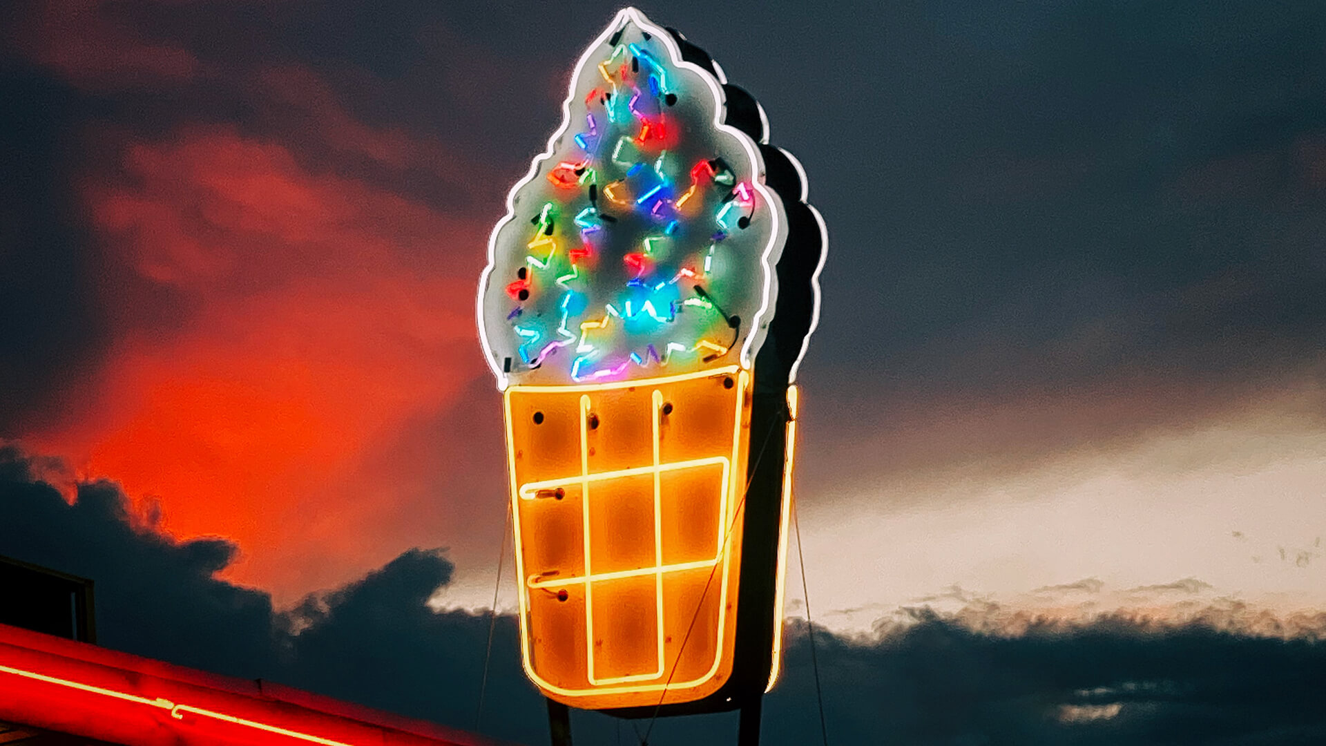 Neon sign of an ice cream cone with sprinkles and dark clouds in the background.
